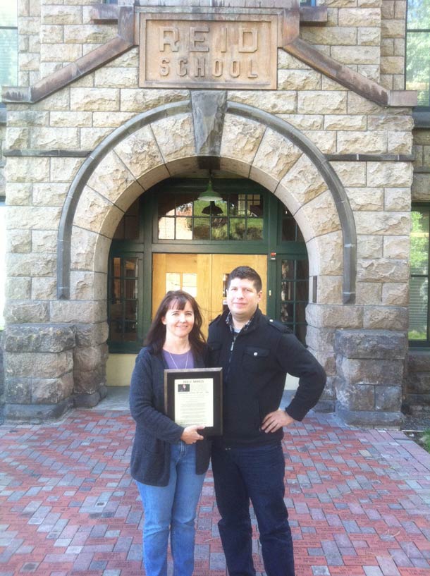 Kelly Cannon-Miller and Kevin Sean Michaels standing in front of a stone building marked Reid School, holding a plaque
